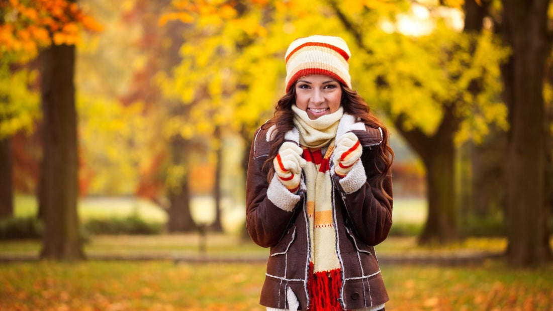 Frau in warmer Herbstkleidung mit Mütze, Schal und Handschuhen steht lächelnd in einem herbstlichen Park mit bunten Blättern und gelb-orangen Bäumen im Hintergrund.
