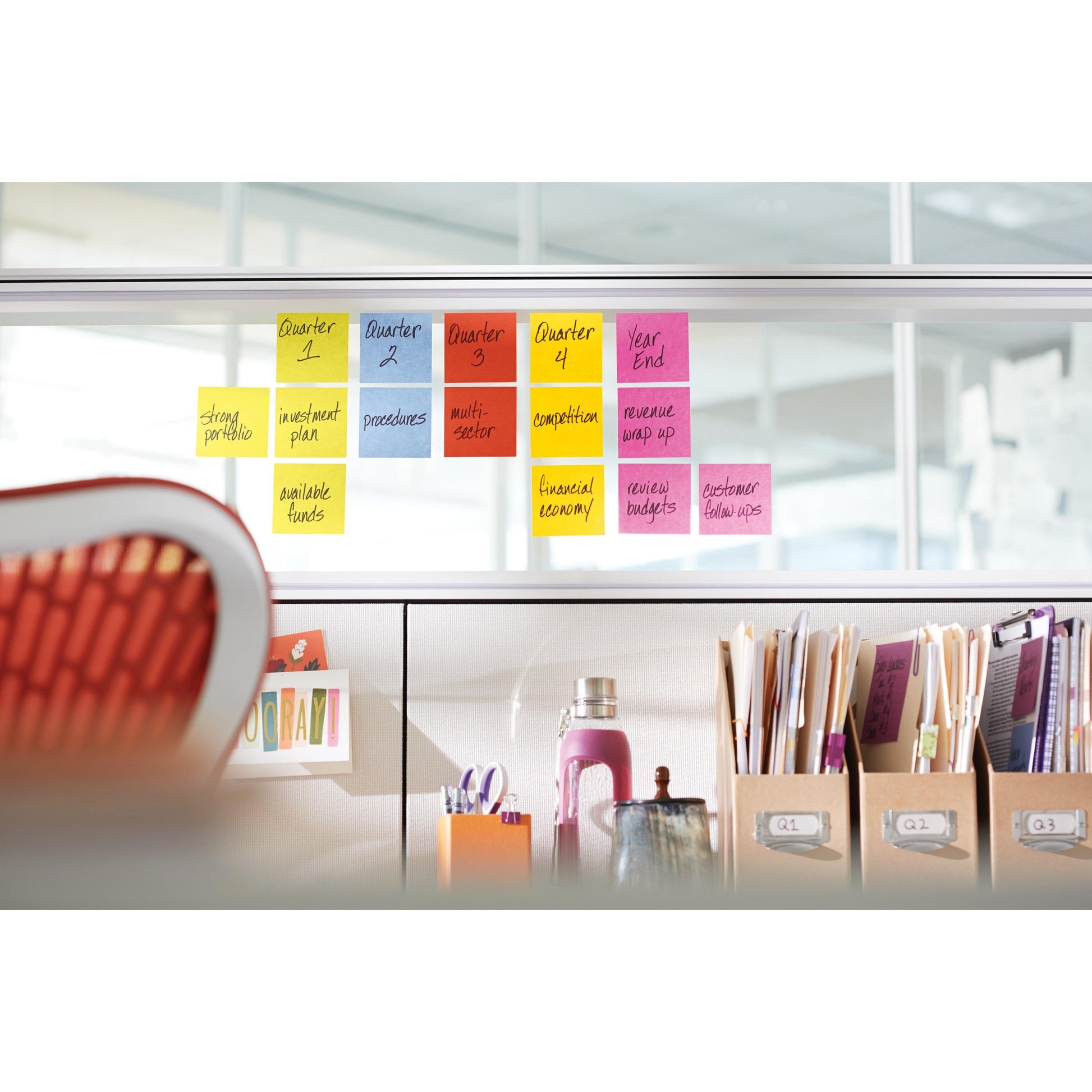Eine Reihe farbenfroher Post-it® Super Sticky Z-Notes (76x76 mm, 90 Blatt/Block, 20+4er Pack) der 3M Deutschland GmbH zeigt Quartalsthemen auf einer Bürotrennwand, darunter geordnetes Büromaterial und die Ecke eines orangefarbenen Stuhls.