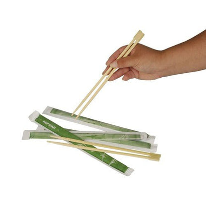 Ruka drží drevené PAPSTAR jedálenské paličky "pure" 23 cm nad niekoľkými pármi, každé jednotlivo zabalené v zelených papierových obaloch. Produkt: PAPSTAR jedálenské paličky "pure", 50 párov | Fólia od PAPSTAR GmbH, zobrazené na bielom pozadí.
