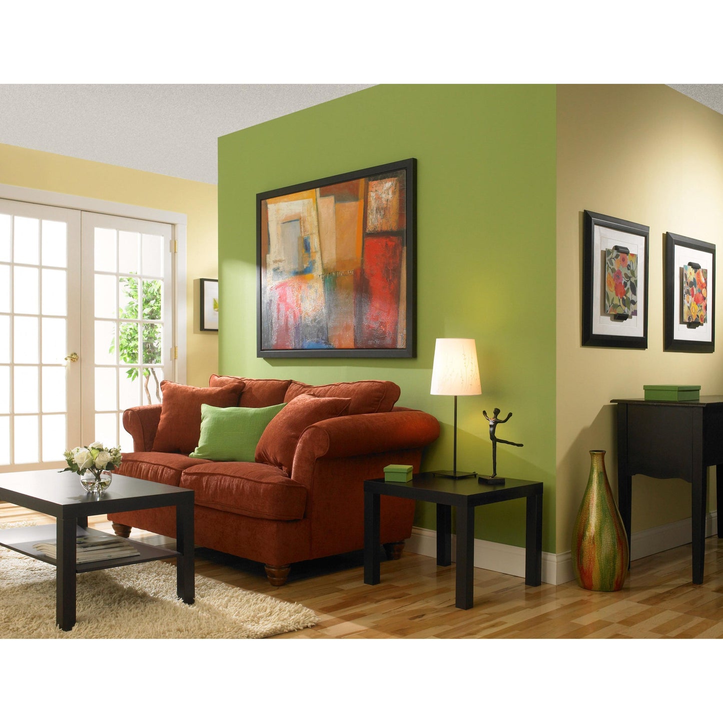Ein Wohnzimmer mit gelben und grünen Wänden, die mit ScotchBlue™ Malerabdeckband "Multi-Surface" von 3M Deutschland GmbH gestrichen sind, einem roten Sofa mit grünen Kissen, einem schwarzen Couchtisch, einem Beistelltisch mit Lampe, abstrakter Kunst, Holzboden und Glastüren.