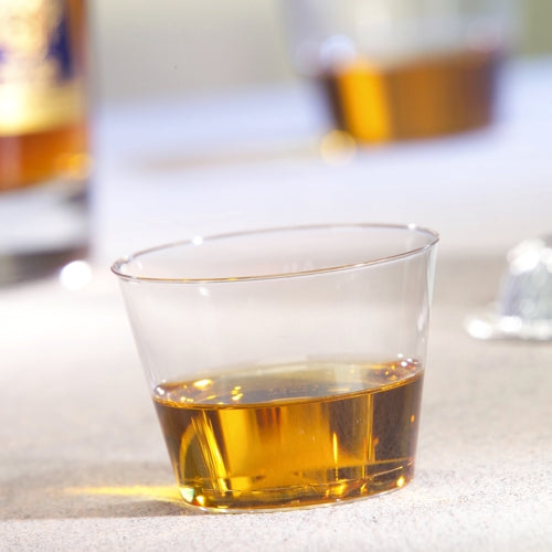 Pohár na pitie Starpak 15, PS 0,1 l krištáľovo čistý, obsahuje malé množstvo jantárovej tekutiny na svetlom povrchu, zatiaľ čo v pozadí je ďalší pohár a fľaša rozmazané.