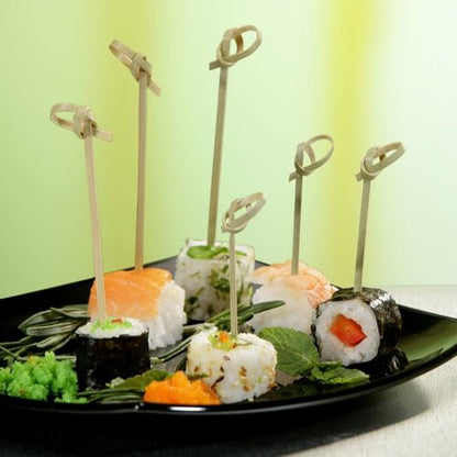 Na čiernom tanieri ležia rôzne sushi rolky, napichnuté každá na PAPSTAR 250 Fingerfood - Picker „pure“ od PAPSTAR GmbH, usporiadané so zelenými ozdobami pred rozmazaným žltozeleným pozadím.