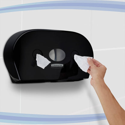 Osoba vyťahuje toaletný papier z čierneho Kimberly-Clark Professional™ dávkovača toaletného papiera - centrálne odoberanie (Jumbo) od Kimberly-Clark GmbH, ktorý je pripevnený na obloženej stene s modrými zakrivenými pruhmi.