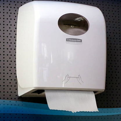 Biely nástenný dávkovač s priehľadným okienkom drží rolku papierových uterákov Scott® (304,00 m x 19,80 cm) od Kimberly-Clark GmbH s visiacim listom na dierovanej šedej stene.