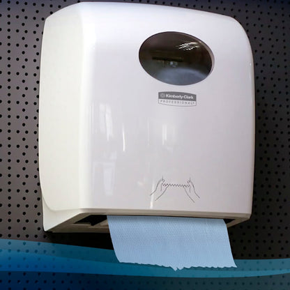 Biely nástenný dávkovač od Kimberly-Clark GmbH drží rolku papierových uterákov Scott® (304,00 m x 19,80 cm) s modrým uterákom a jednoduchým ilustrovaným návodom pred tmavým pozadím z dierovaného plechu.