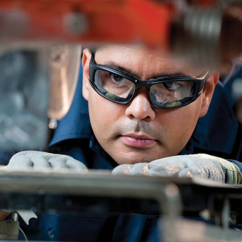 Ein Mann trägt Kimberly-Clark GmbH Auslaufartikel: KleenGuard® V50 Calico™ Schutzbrille mit Beschlagfrei-Beschichtung und Handschuhen prüft konzentriert ein Maschinenteil im Industriebereich - für klare Sicht und maximale Sicherheit.