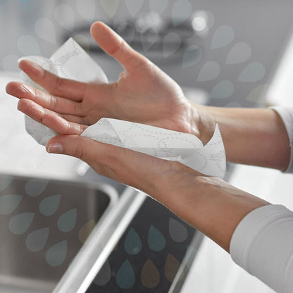 Osoba si utiera ľavú ruku bielou Kleenex® Ultra™ rolkovou utierkou (2-vrstvová, E-Rolka Large) od Kimberly-Clark GmbH nad umývadlom na hygienické sušenie. Grafika s kvapkami vody prekrýva obraz.