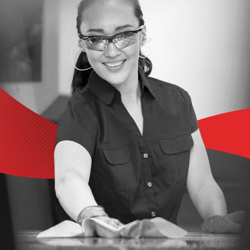 Žena s rukavicami a okuliarmi sa usmieva, zatiaľ čo čistí povrch utierkou WypAll® X60 General Clean - BRAG™ Box (41,80 × 28,30 cm) od spoločnosti Kimberly-Clark GmbH; v pozadí sa objavuje červený grafický dizajn.