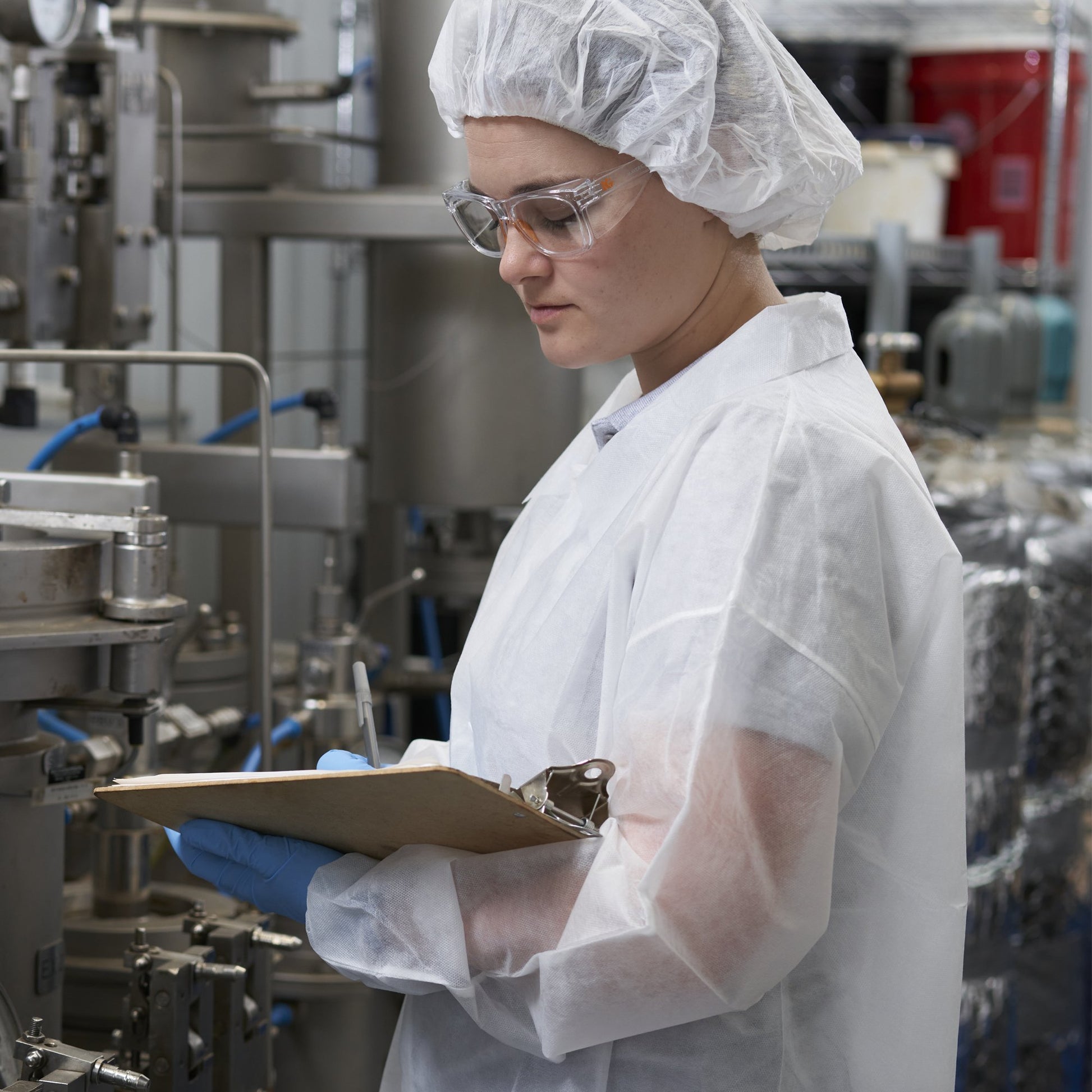 Eine Person mit Haarnetz, Schutzbrille und blauen Handschuhen trägt einen weißen KleenGuard® KGA10 Leichter Schutzkittel von Kimberly-Clark GmbH und schreibt auf einem Klemmbrett in einem Industrielabor mit verschiedenen Arbeitsumgebungen im Hintergrund.