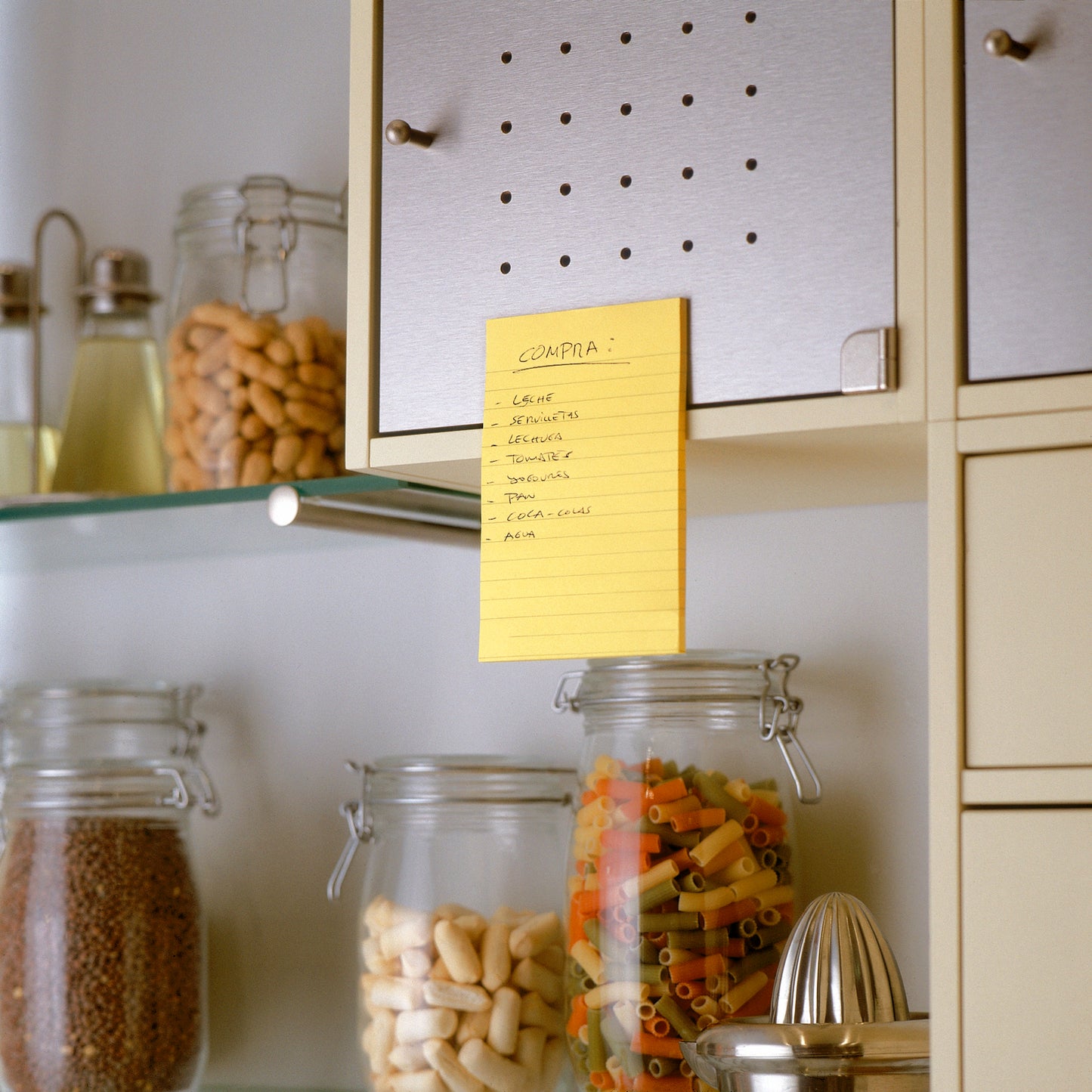 Ručne napísaný nákupný zoznam v španielskom jazyku sa nachádza na béžovej kuchynskej skrinke pomocou 3M Deutschland GmbH Post-it® Super Sticky Notes vo veľkom formáte, Ultra Žltá (101 x 152 mm), nad sklenenými nádobami s cestovinami, fazuľami a obilninami. V susedných policiach sa nachádzajú ďalšie poháre a kuchynské náčinie.