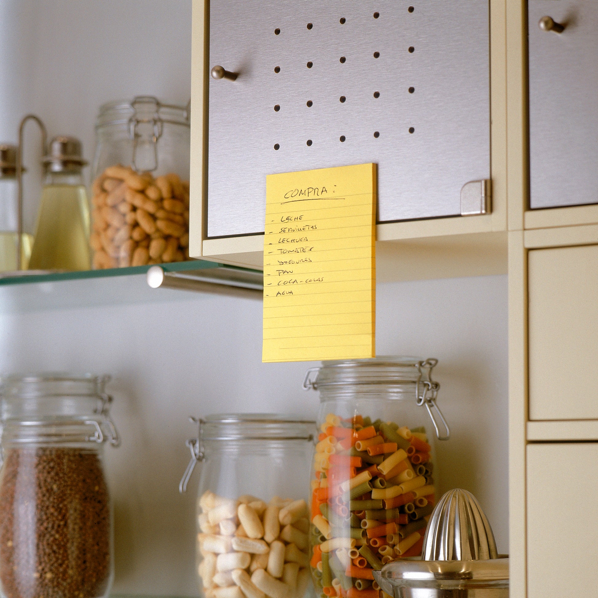 Ein Post-it® Super Sticky Note in Ultra Gelb (101x152mm, 3M Deutschland GmbH) ist an einem Küchenschrank über Gläsern mit Nudeln, Mandeln und mehr angebracht. Öl- und Essigflaschen stehen daneben auf einem Glasregal.