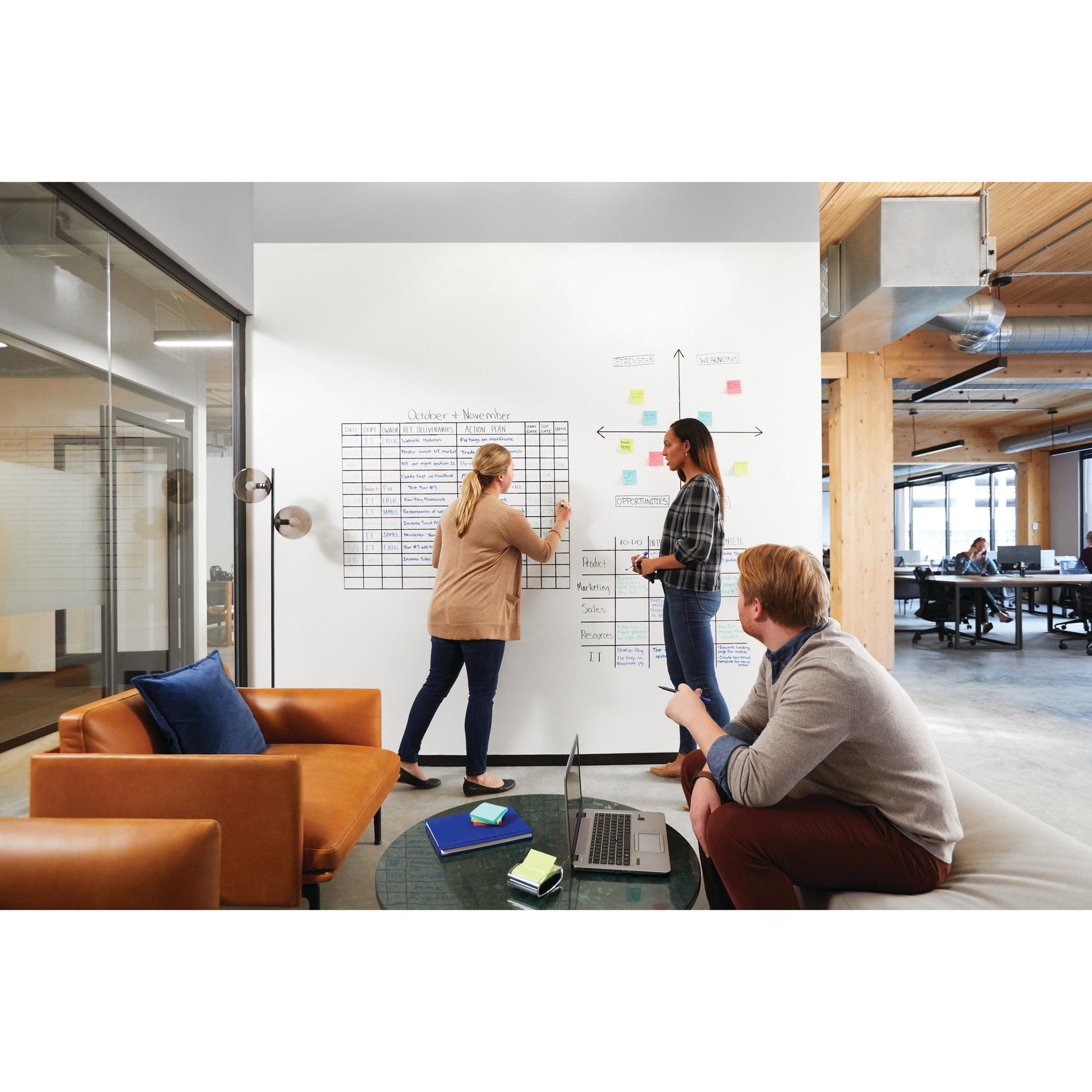 Drei Personen sitzen in einem modernen Büro. Zwei benutzen die Post-it® Easy Erase Whiteboard-Oberfläche, Weiß von 3M Deutschland GmbH, und fügen Charts und Haftnotizen hinzu; einer sitzt auf einer Couch mit einem Laptop. Große Fenster und ein offener Arbeitsbereich sind zu sehen.