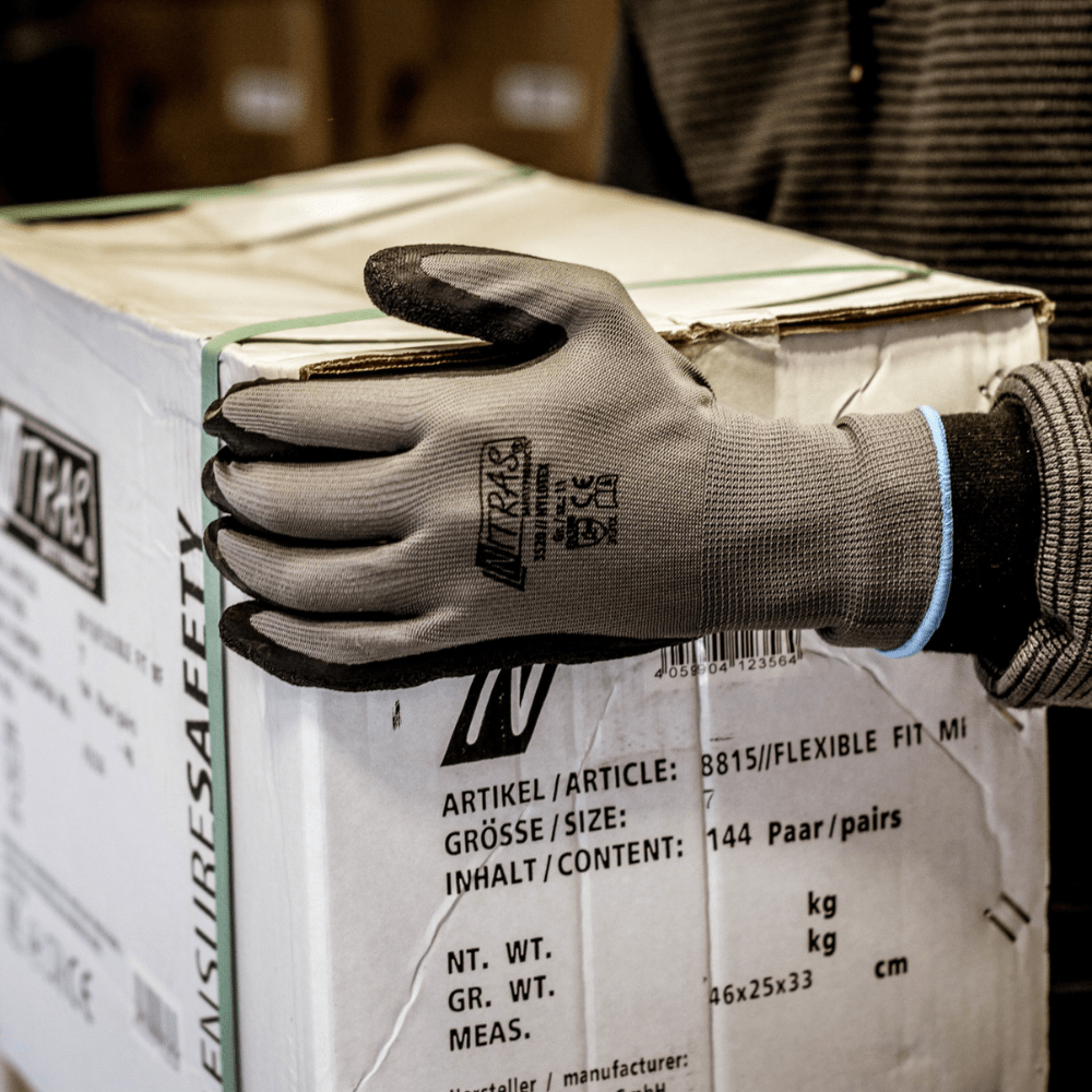 Osoba, ktorá nosí nylonové rukavice Nitras Nylotex s latexovým povlakom od AS Arbeitsschutz GmbH, drží v ruke veľkú bielu škatuľu s nemeckým a anglickým popisom; viditeľná je len jej horná časť tela a čiastočne ruka.