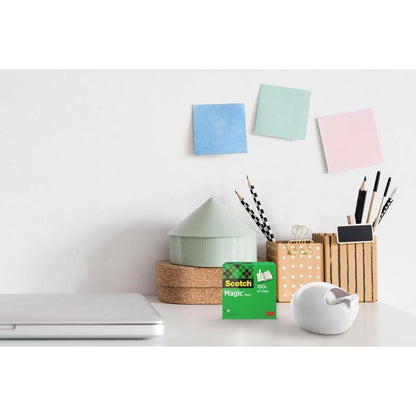 Stôl s uzavretým laptopom, stolový držiak pásky Scotch™ Karim Rashid od 3M Deutschland GmbH a škatuľka s lepiacou páskou, nádoba na perá a ceruzky, malé ozdoby v tvare domu a tri farebné samolepiace lístky na bielej stene.