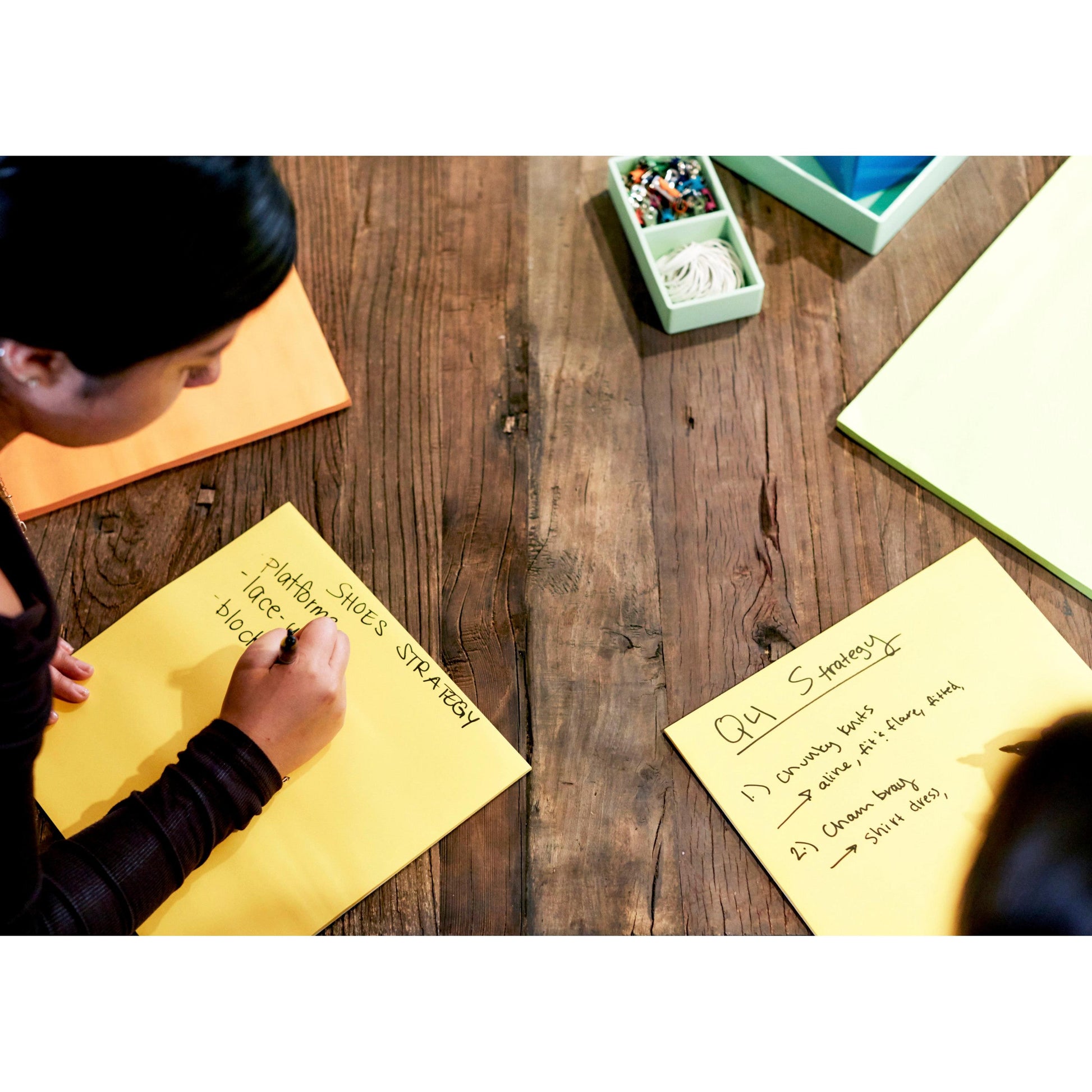 Jemand schreibt auf einen gelben Post-it® Super Sticky Big Note BN11-EU der 3M Deutschland GmbH mit der Aufschrift "Platform Shoes Strategy", während auf einem anderen Zettel in der Nähe "Q4 Strategy" steht. Beide Zettel liegen zusammen mit Büromaterial auf einem Holztisch.