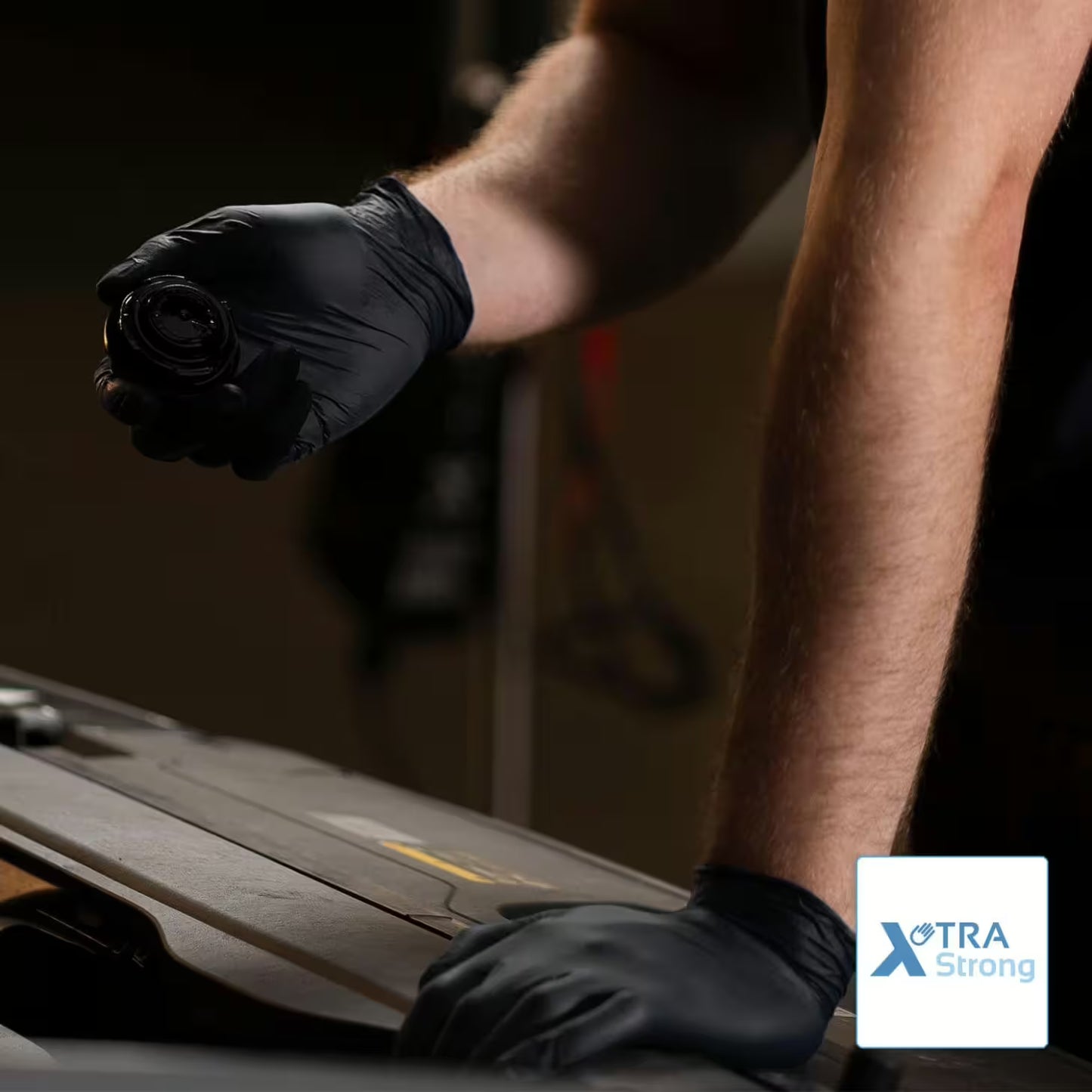 Osoba, ktorá nosí jednorazové rukavice ARNOMED NITRIL XTRA PURE BLACK od spoločnosti ARNOWA GmbH, drží okrúhlu súčiastku auta nad otvoreným motorom, ruka spočíva na ňom. V jednom rohu je logo "XTRA Strong".
