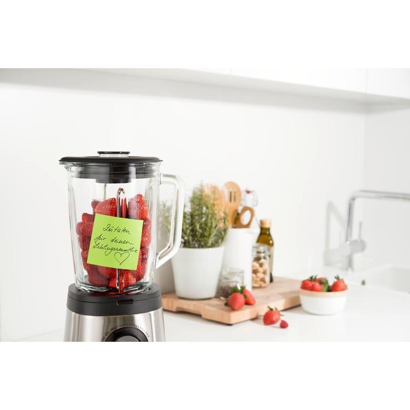 Mixér s jahodami stojí na bielej kuchynskej linke. Post-it® Super Sticky Note (76 x 76 mm, 90 listov, 100 % PEFC) od 3M Deutschland GmbH je pripevnený na nádobe. V blízkosti sa nachádzajú pomôcky, črepníkové rastliny, olivový olej a ďalšie jahody na doske na krájanie.