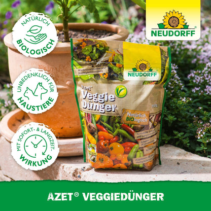 Vrecko Neudorff Shop Azet VeggieDünger stojí na drevenom povrchu vonku vedľa terakotového kvetináča so zelenými rastlinami. Na obale sú zobrazené obrázky zeleniny a certifikačné logá.