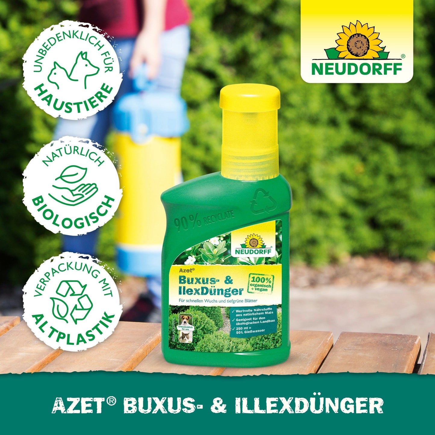 Zelená fľaša od Neudorff Shop Azet Buxus- & Ilex hnojivo tekuté stojí na drevenej ploche vonku. Ikony ukazujú bezpečnosť pre domáce zvieratá, biologické zložky a recyklované obaly. Logo Neudorff sa objavuje v pravom hornom rohu.