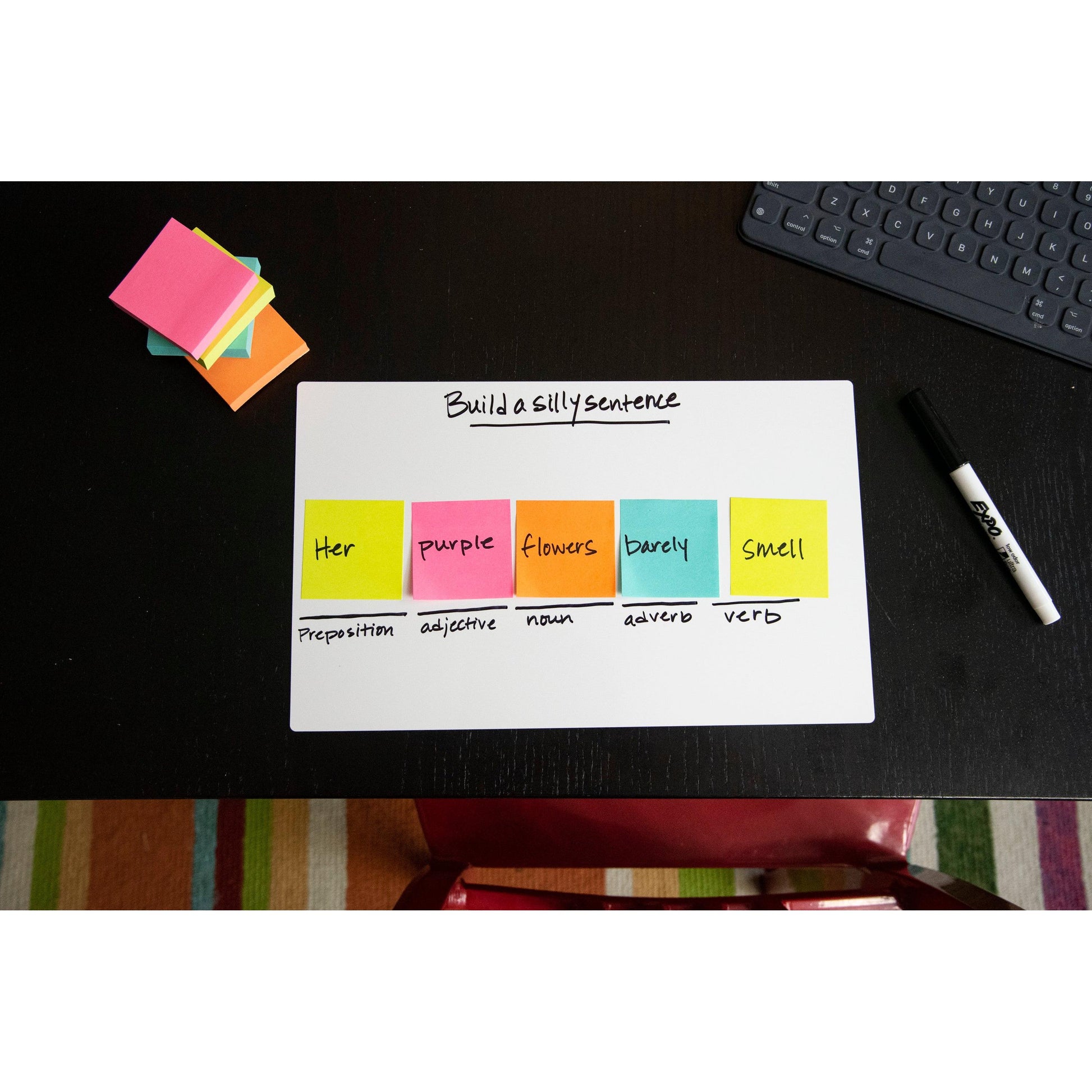 Auf einem dunklen Schreibtisch wird mit bunten 3M Deutschland GmbH Post-it® Super Sticky Notes (76 mm x 76 mm, 90 Blatt/Block), die jeweils nach Wortarten beschriftet sind, ein Satz gebildet. Ein Marker, Haftnotizblöcke und eine Tastatur liegen daneben.
