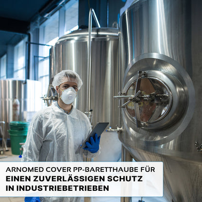 Eine Person in Schutzkleidung, darunter die ARNOMED COVER PP-BARETTHAUBE der ARNOWA GmbH und eine Maske, hält eine Tablette neben Edelstahltanks in einem hygienischen Industriebereich. Der deutsche Text über zuverlässigen Schutz ist unten eingeblendet.