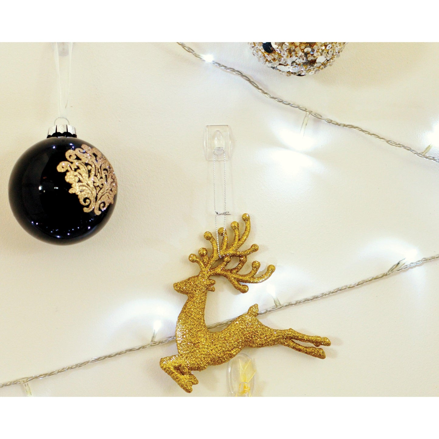 Ein goldenes Glitzer-Rentier-Ornament hängt an einer weißen Wand, sicher befestigt mit einem transparenten Command™ Mini-Haken, 17006CLR von 3M Deutschland GmbH, neben einem schwarzen Ornament mit goldenen Details und weißen Lichterketten.