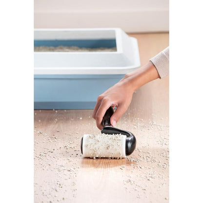 Ruka zbiera so Scotch-Brite™ Everyday Clean Fusselroller na ploché povrchy (56 listov, 1/balenie) od 3M Deutschland GmbH rozsypaný mačací piesok z drevenej podlahy, v pozadí je vidieť mačaciu toaletu s modrým vrchnákom.