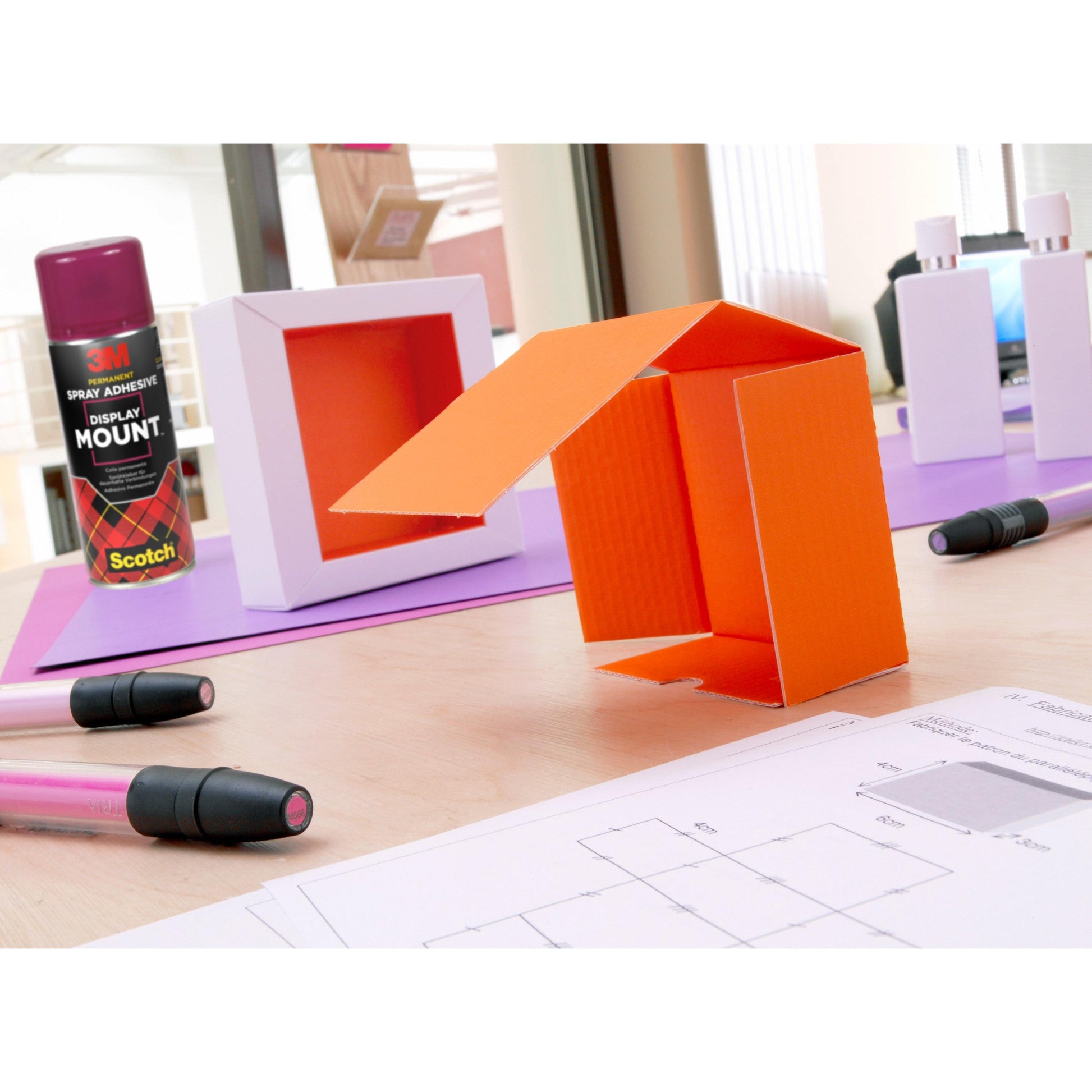 Ein orangefarbenes Architekturmodell aus Pappe steht auf einem Schreibtisch mit Zeichnungen, Markern, weißen Flaschen und einer Dose 3M™ Scotch® DisplayMount™ Klebespray (400 ml, 1 Dose/Packung) der 3M Deutschland GmbH im Hintergrund.