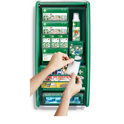 Osoba drží obväz s gélom na popáleniny pred stanicou prvej pomoci a popálenín Cederroth (1 balenie) od Orkla Wound Care AB, zelenou stanicou prvej pomoci s materiálmi na popáleniny, obväzmi, fľašami a návodmi.