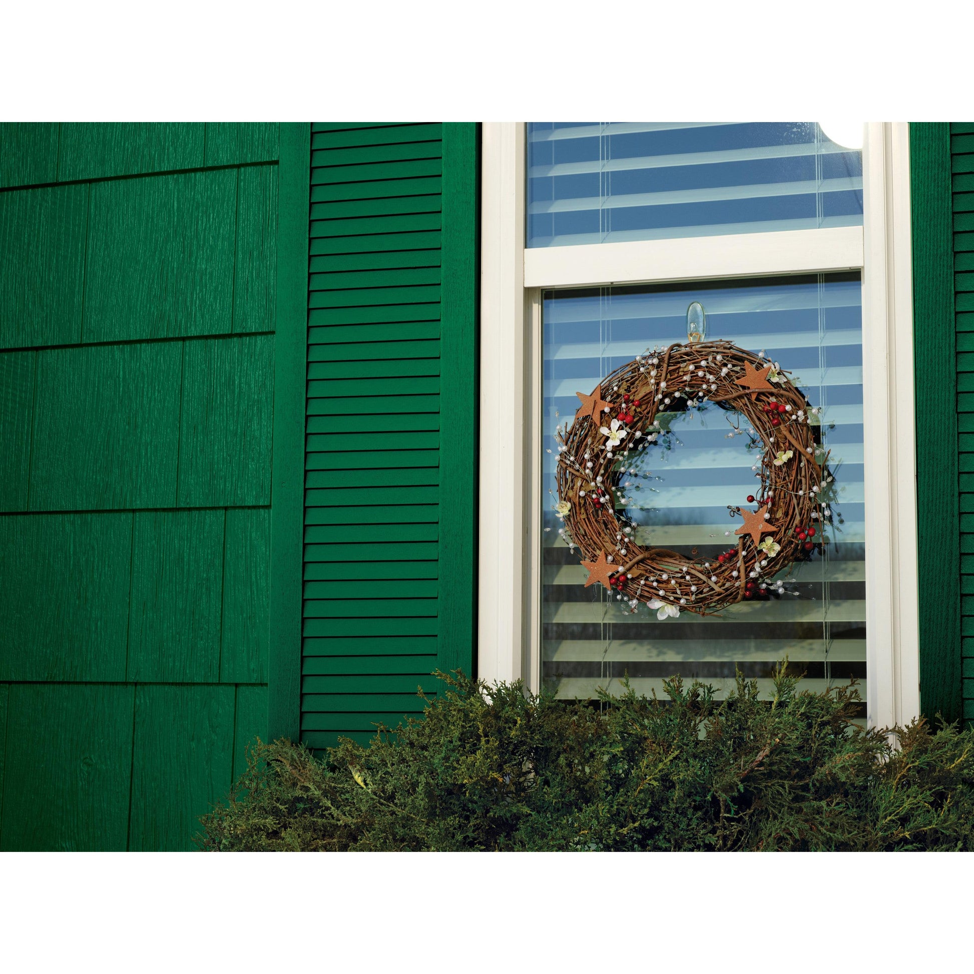 In einem Fenster mit weißen Jalousien hängt ein dekorativer Kranz, der mit einem transparenten mittelgroßen Command™ Haken M für den Außenbereich von 3M Deutschland GmbH angebracht wurde; das Haus hat eine grüne Fassade und Fensterläden, unter dem Fenster befinden sich Büsche.