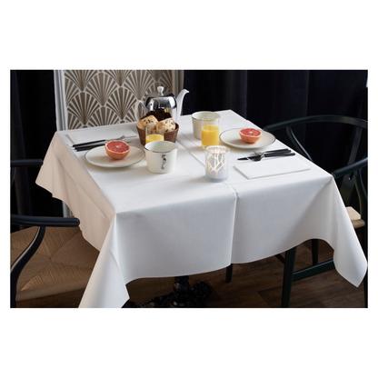 Raňajkový stôl je elegantne prestretý s bielym obrusom, dvoma stoličkami a vázou s geometrickým vzorom. Na stole sú polovice grapefruitu, pečivo, čaj a pomarančový džús. Duni Dunilin®-servítka (48x48 cm) od Duni GmbH dodáva šarm a funkčnosť atmosférickému rannému prestieraniu.