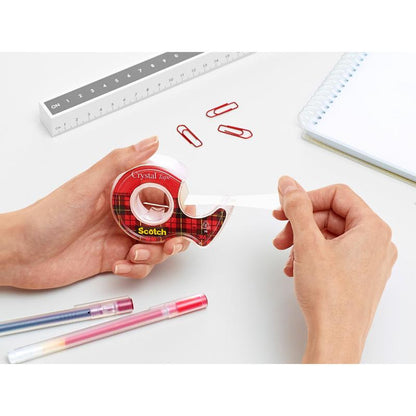 Osoba vyťahuje lepiacu pásku Scotch® Crystal (19 mm x 7,5 m) z červeného ručného odvíjača od 3M Deutschland GmbH na bielom stole s červenými sponkami, perami, pravítkom a špirálovým zápisníkom.