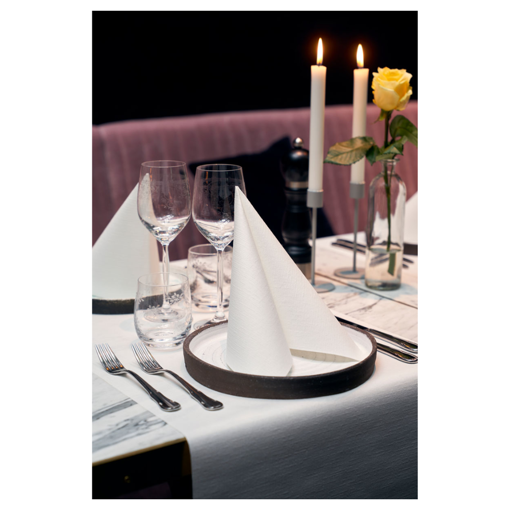 Jedálenský stôl je elegantne prestretý dvoma Duni Dunilin®-servítkami 48 x 48 cm biela, ktoré sú zložené do trojuholníkov na tanieroch. Dve veľké horiace sviečky a žltá ruža vo váze zdobia pozadie. Poháre na víno, pohár na vodu a príbor dopĺňajú túto sofistikovanú atmosféru s látkovým šarmom.