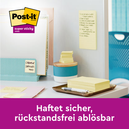 Uprataný písací stôl s 3M Deutschland GmbH Post-it® Super Sticky Notes, Žltá, 47,6 mm x 47,6 mm (PEFC-certifikovaný), pripevnený na deliacu stenu. Nemecký text: "Spoľahlivo drží, bez zvyškov odstrániteľný." Stohy poznámok a písacie potreby na stole.