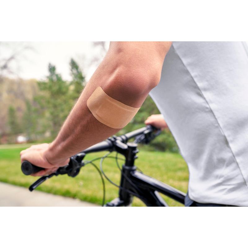 Osoba, ktorá nosí bielu košeľu a jazdí na bicykli vonku, má na odhalenom ramene Nexcare™ Ultra Stretch MAXI Comfort Flexible náplasť (50 mm x 101 mm) od 3M Deutschland GmbH; v pozadí je vidieť zelenú trávu a stromy.