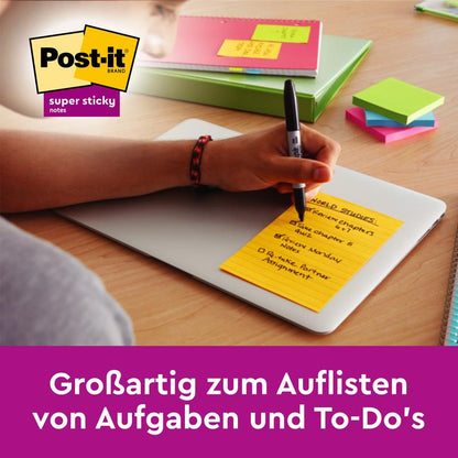 Ruka píše na ružový veľkoformátový Post-it® Super Sticky Notes od 3M Deutschland GmbH, linajkovaný (127 x 203 mm), na zatvorenom notebooku. Vedľa ležia ďalšie lepiace poznámky a perá. Perfektné na písanie zoznamov a plánovanie úloh.