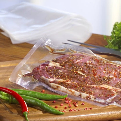 Starpak 100 zatavovacie vrecko (PA/PE priehľadné), naplnené marinovaným surovým mäsom, leží na drevenej doske, obklopené zelenými a červenými chilli papričkami; v pozadí sú rozmazané petržlen a kuchynské potreby.