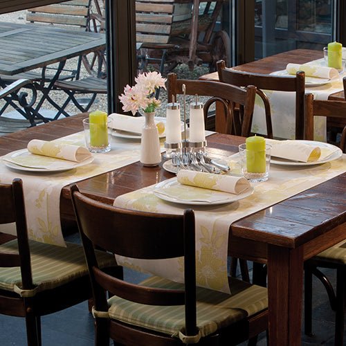 Drevený jedálenský stôl pre štyri osoby s bielymi taniermi, PAPSTAR 50 obrúskov "ROYAL Collection" 1/4-sklad 40 x 40 cm zelené od PAPSTAR GmbH, zelenými sviečkami, sklom, soľničkou a koreničkou a vázou s ružovými kvetmi pri okne.