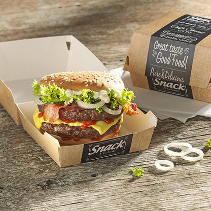 Cheeseburger so šalátom, paradajkou, slaninou, cibuľou a omáčkou leží v otvorenej PAPSTAR 50 burgerovej krabici z kartónu "pure" od PAPSTAR GmbH na drevenom stole. Vedľa je zatvorená krabica s nápisom "Good Food" a cibuľovými krúžkami.