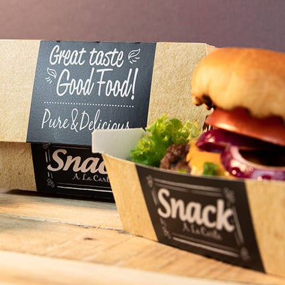Kartónový podnos s nápisom "Snack A La Carte" obsahuje hamburger so šalátom, paradajkou a cibuľou. V pozadí je PAPSTAR 50 burgerových krabíc, kartón "pure" "Good Food" od PAPSTAR GmbH: Dobrý vkus Good Food! Čisté a chutné.