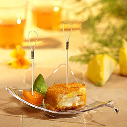 Sklenená misa predstavuje obaľovanú, vyprážanú predjedlovú kocku s PAPSTAR Fingerfood Picker "Loop" (10 cm), segmentom pomaranča a zelenou ozdobou. V pozadí sa rozmazávajú citrónové kliny a dva nápoje spoločnosti PAPSTAR GmbH.