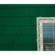 Eine Nahaufnahme einer grünen Schindelwand mit einem weiß gerahmten Fenster auf der rechten Seite. 3M Command™ Outdoor Lichter-Clips (17017CLR-AWPU) befestigen weiße Lichterketten sicher um den Rahmen, während horizontale Jalousien das Fenster abdecken.