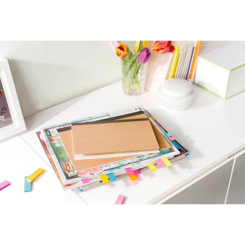 Stoh kníh a hnedý zápisník s farebnými 3M Post-it® Page Markermi z Poptimistic Collection ležia na bielom stole. V pozadí sú tulipány vo váze, ďalšie knihy a biely okrúhly objekt.