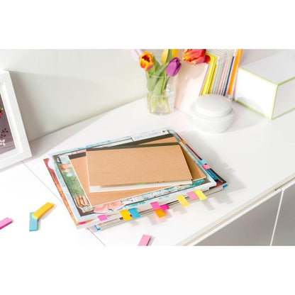 Stoh kníh a hnedý zápisník s farebnými 3M Post-it® Page Markermi z Poptimistic Collection ležia na bielom stole. V pozadí sú tulipány vo váze, ďalšie knihy a biely okrúhly objekt.