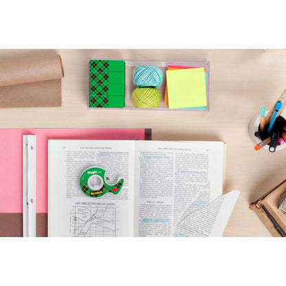 Pracovný priestor s otvorenou knihou, milimetrovým papierom, farebnými klbkami nite, poznámkovými lístkami, perami v pohári, gumami, rolkou hnedého baliaceho papiera a rolkou 3M Deutschland GmbH Scotch® Magic™ Neviditeľná lepiaca páska na svetlom drevenom stole.