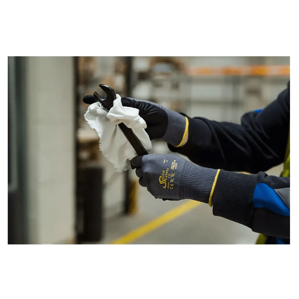 Osoba s ultraľahkými montážnymi rukavicami AMPri SolidSafety Tough Air od AMPri Handelsgesellschaft mbH opatrne čistí skrutkový kľúč bielou handrou v interiéri. Sivé rukavice, ktoré sú ocenené úrovňou odolnosti proti oderu 4, vynikajú na rozmazanom pozadí priemyselných regálov a zariadení.