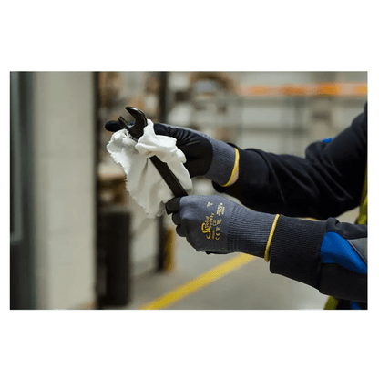 Osoba s ultraľahkými montážnymi rukavicami AMPri SolidSafety Tough Air od AMPri Handelsgesellschaft mbH opatrne čistí skrutkový kľúč bielou handrou v interiéri. Sivé rukavice, ktoré sú ocenené úrovňou odolnosti proti oderu 4, vynikajú na rozmazanom pozadí priemyselných regálov a zariadení.