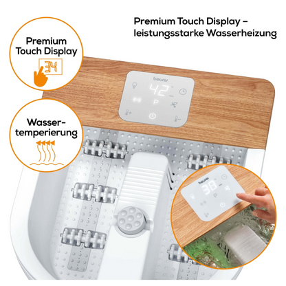 Kúpeľ na nohy značky Beurer FB 65 Wellness vírivý kúpeľ na nohy od Beurer GmbH má biely základ a drevenú dosku, s digitálnym dotykovým displejom, ktorý zobrazuje teplotu 42 °C. Na etiketách je napísané „Ohrievanie vody“ a „Prémiový dotykový displej“. Ruka nastavuje displej a masážne valčeky pre vibračnú a bublinkovú masáž sú viditeľné.