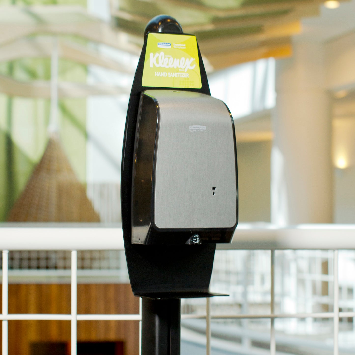 Ein Kimberly-Clark GmbH Elektronischer Waschlotionen Ständer (Schwarz, 1 Spender) ist innen nahe einem weißen Metallgeländer für hygienische Handreinigung aufgestellt, unscharfe architektonische Details im Hintergrund.