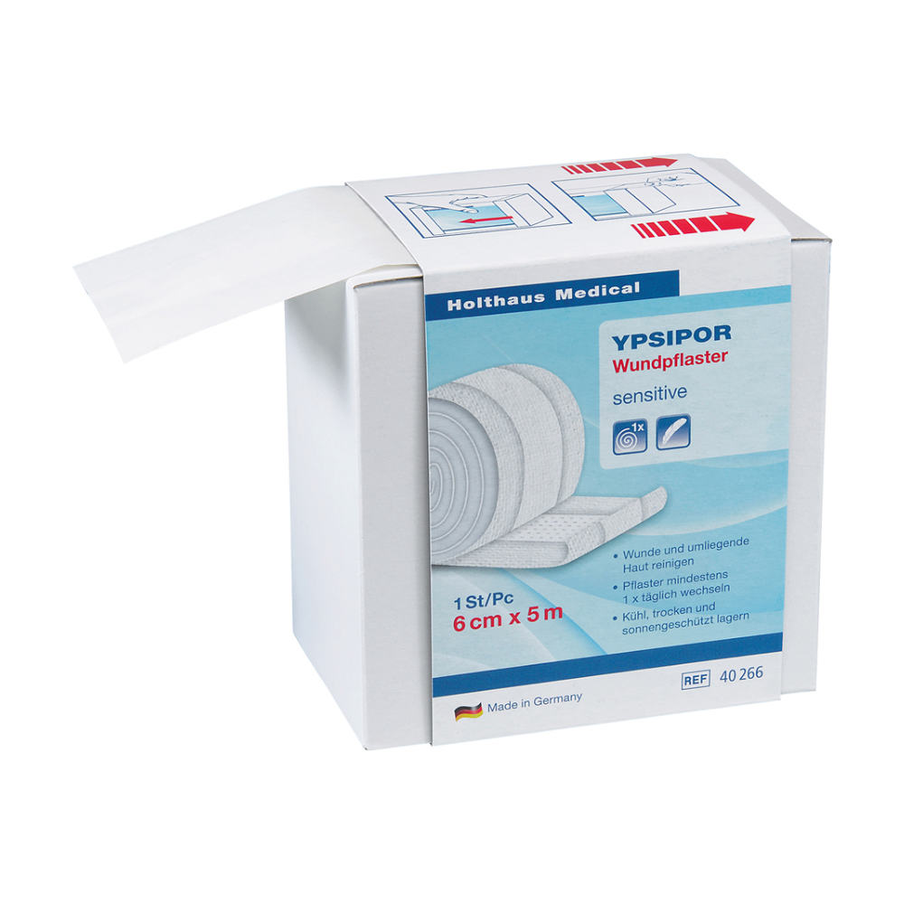Zobrazená je krabička Holthaus Ypsipor Wundpflaster Vlies od Holthaus Medical GmbH & Co. KG. Na krabičke je uvedené, že je citlivá, má rozmery 6 cm x 5 m a je vyrobená v Nemecku. Časť pokožke priateľskej vliesovej obväzovej rolky je viditeľná a vyčnieva z krabičky.